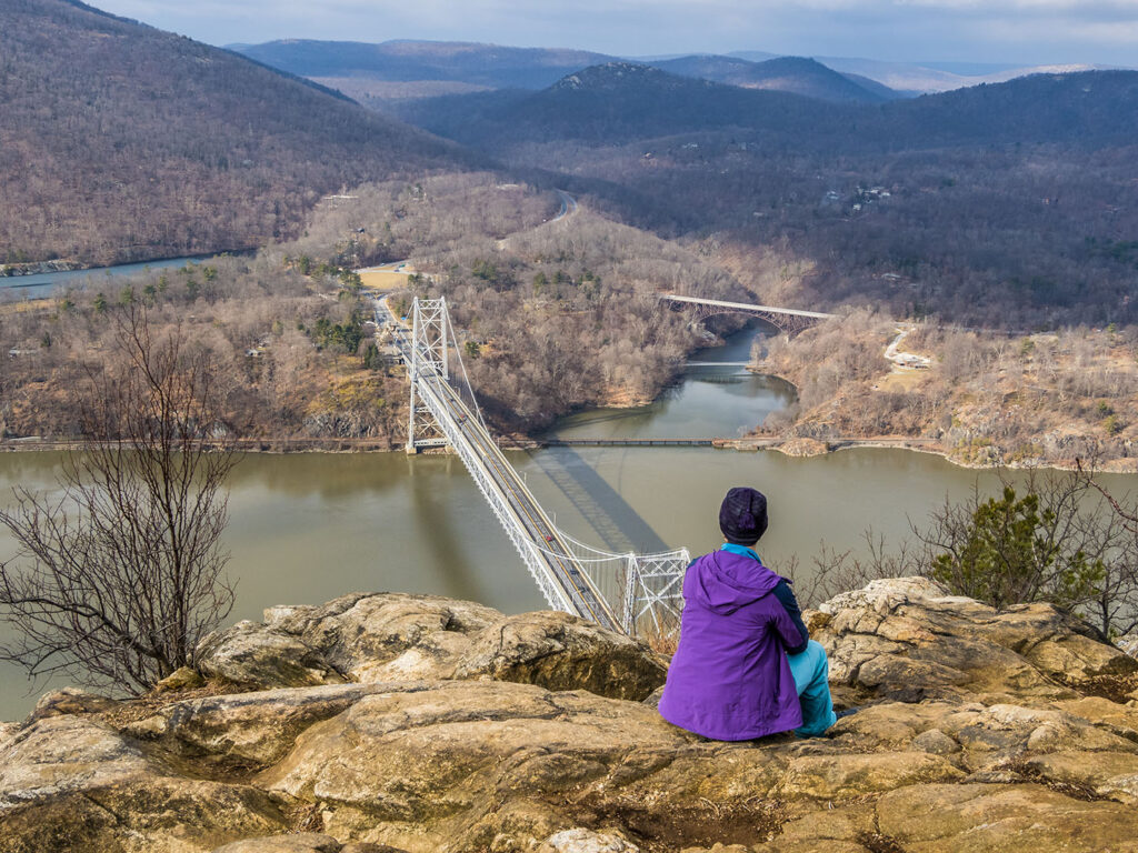 Anthony’s Nose Overlook: A Stunning Hudson Valley View of Bear Mountain Bridge - Hudson Valley Luxury Resorts A hiker in a purple jacket sits on the Anthony’s Nose summit, overlooking Bear Mountain Bridge and the Hudson River in the Hudson Valley.