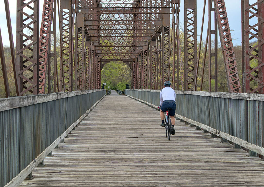 Cycling Across a Historic Bridge on the Dutchess Rail Trail - Hudson Valley Luxury Resorts A cyclist in a white jersey rides across a wooden walkway on a historic railroad bridge along the Dutchess Rail Trail.