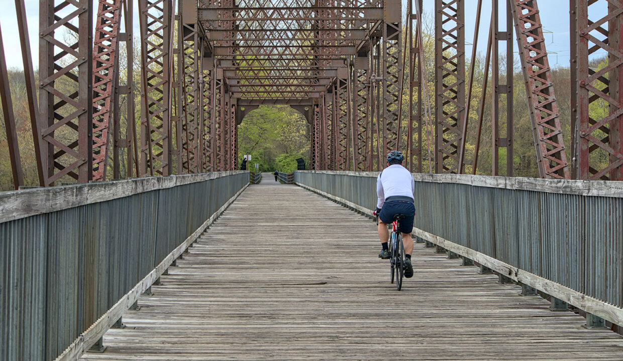 A cyclist in a white jersey rides across a wooden walkway on a historic railroad bridge along the Dutchess Rail Trail.