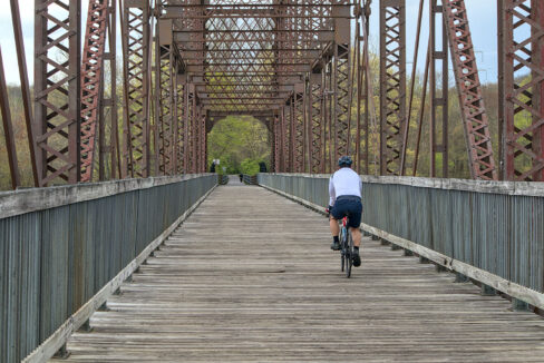 A cyclist in a white jersey rides across a wooden walkway on a historic railroad bridge along the Dutchess Rail Trail.