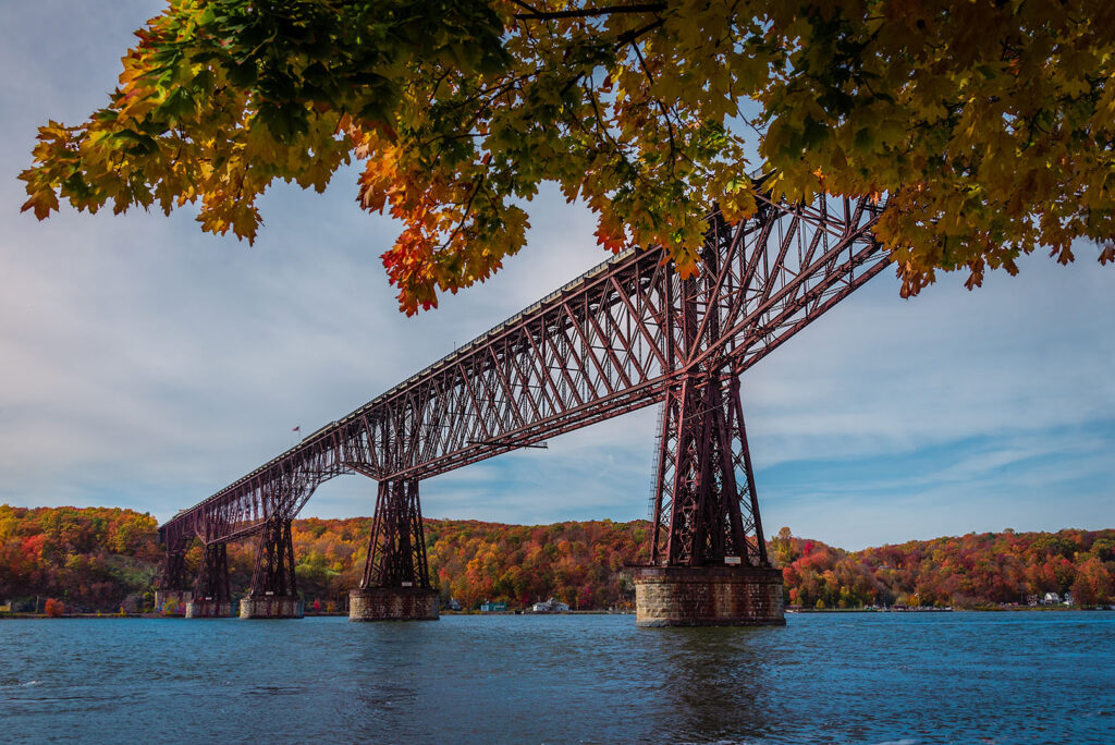 Autumn Splendor on the Dutchess Rail Trail Bridge - Hudson Valley Luxury Resorts An old iron railroad bridge spanning the Hudson River, framed by vibrant autumn leaves along the Dutchess Rail Trail.