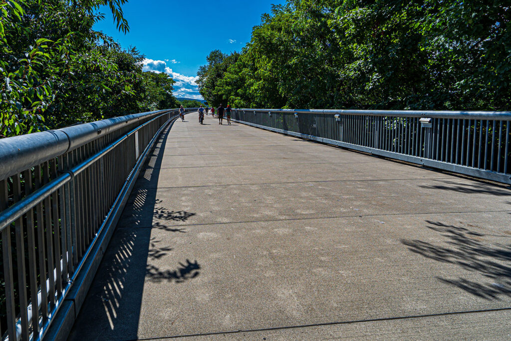 A Picturesque Stroll: Bicyclists and Pedestrians on the Walkway Over the Hudson - Hudson Valley Luxury Resorts A wide concrete walkway on the Walkway Over the Hudson, with railings on both sides and people biking and walking under a bright blue sky.