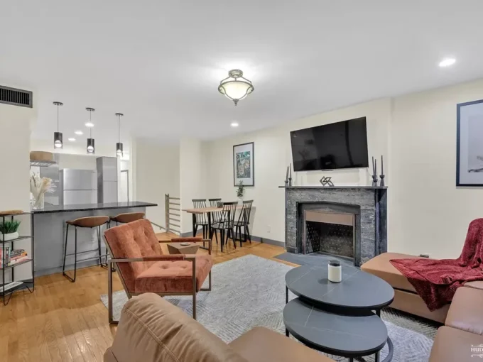 Open plan living room with tan leather chair, gray sofa, stone fireplace, mounted TV, and kitchen breakfast bar with pendant lights