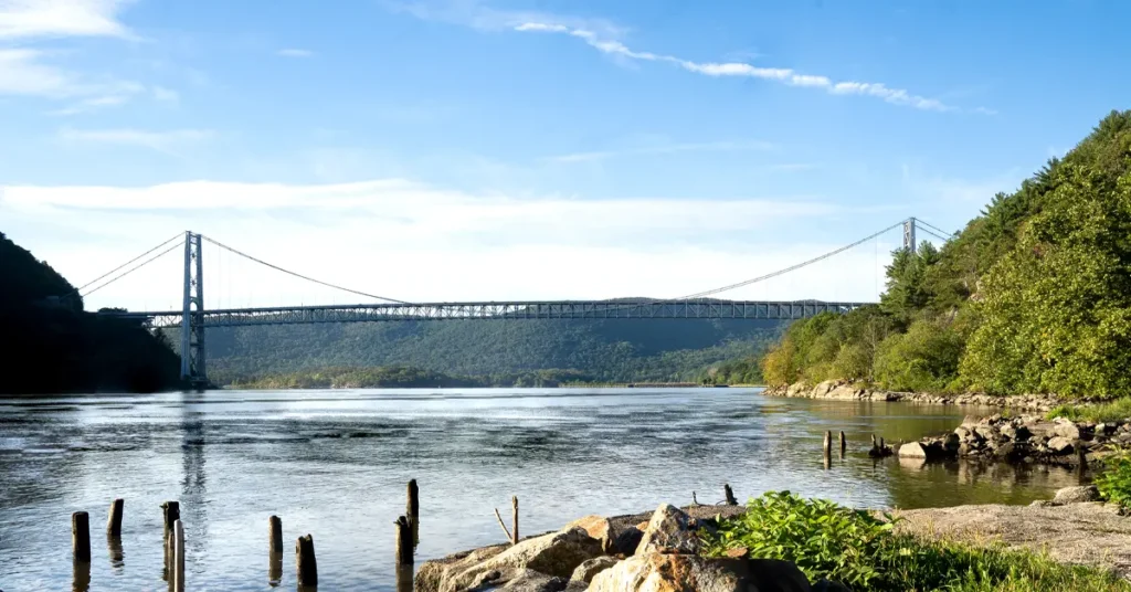 Bear Mountain Bridge suspension span crossing the Hudson River viewed from rocky shoreline with weathered wooden pilings in foreground, forested hills on both banks under blue sky with wispy clouds