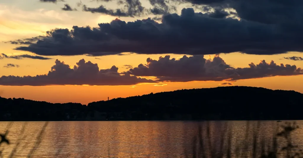 Dramatic sunset over the Hudson River with vivid orange and gold sky, dark storm clouds, silhouetted hills along horizon, golden light reflecting on calm water, and reed grasses in foreground
