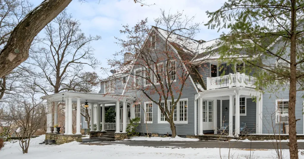 Large gray-shingled Colonial-style farmhouse with white columned portico entrance, second-floor balcony with white railings, and stone steps surrounded by bare winter trees with light snow covering the ground