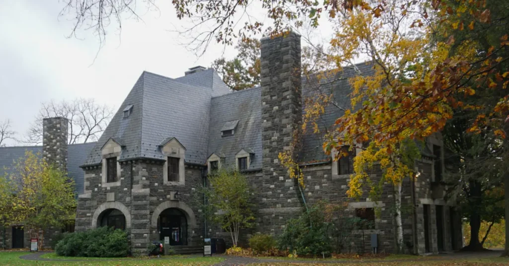 Gray stone castle-like building with steep slate roof, tall stone chimneys, arched entrance portal, and dormer windows surrounded by trees displaying golden fall foliage on overcast autumn day
