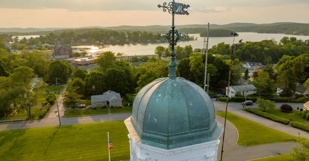 Aerial view of weathered verdigris copper dome with ornate iron weathervane overlooking Lake Mahopac and tree-lined village streets at golden hour with distant wooded hills in Putnam County