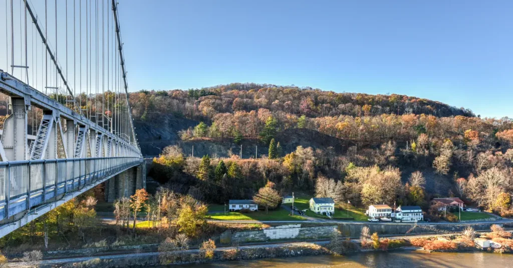 Steel suspension bridge spanning the Hudson River with late autumn foliage on steep hillside, small riverside homes and railroad tracks along the riverbank under clear blue sky